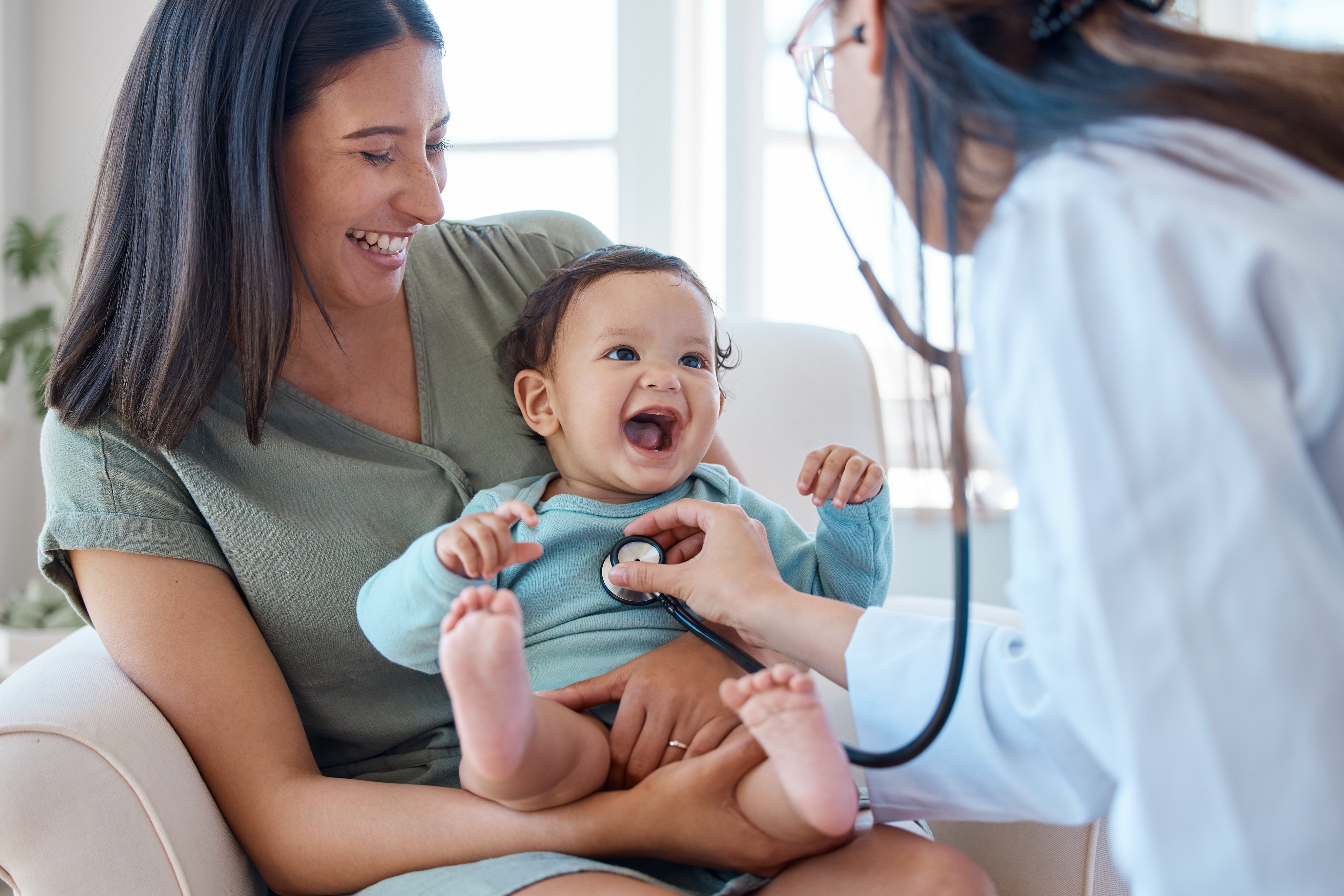 Woman holding smiling baby while the baby is being inspected by a doctor